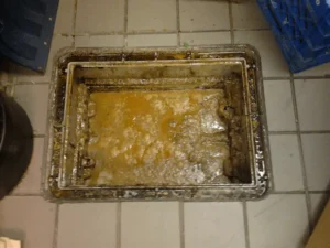 Caixa de gordura retangular aberta e completamente entupida com gordura petrificada em piso de cerâmica.