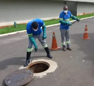 Dois técnicos de desentupidora uniformizados realizando manutenção e desobstrução de rede de esgoto externa em bueiro na rua.